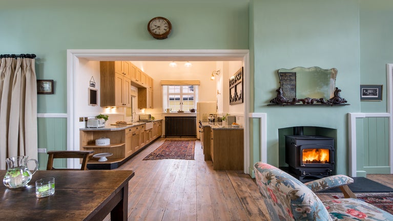 The open plan dining area, sitting area and kitchen at Crabtree School House, Devon. A section of the wooden dining table, the floral seat in the sitting area and the kitchen.